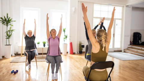 People in class of chair yoga with hands stretched above their heads while in chair. 