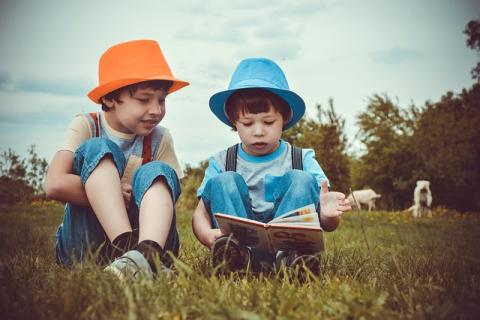 Two little boys reading together in field, one with orange hat the other with blue hat. 